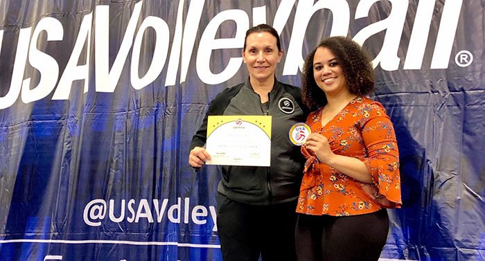 Two women stand in front of a USA Volleyball banner with one holding a certificate and the other holding a medal