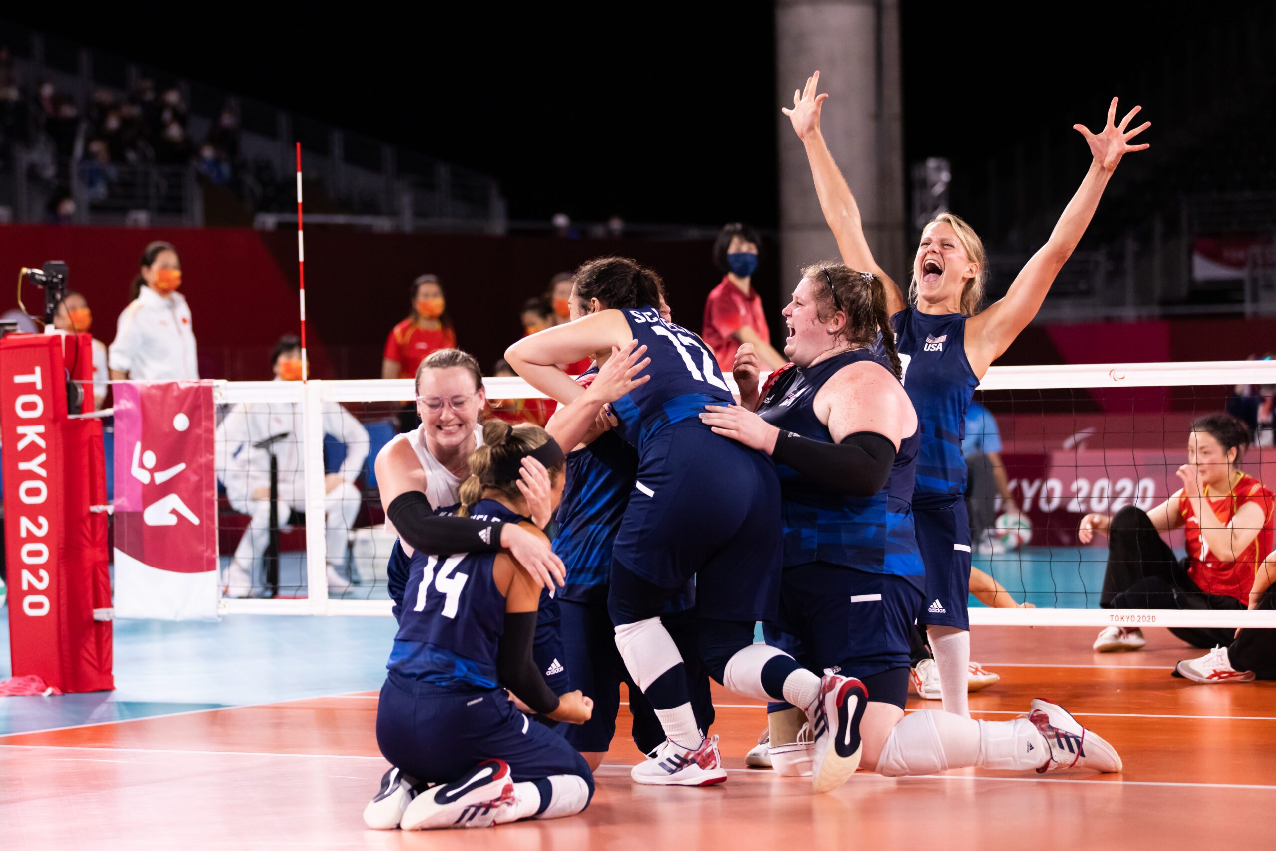 The U.S. women's sitting national team celebrates and hugs on a volleyball court in front of a net.