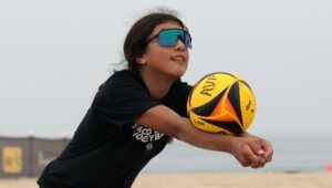 An athlete passes a volleyball with her forearms. She is wearing sunglasses and is on a beach.