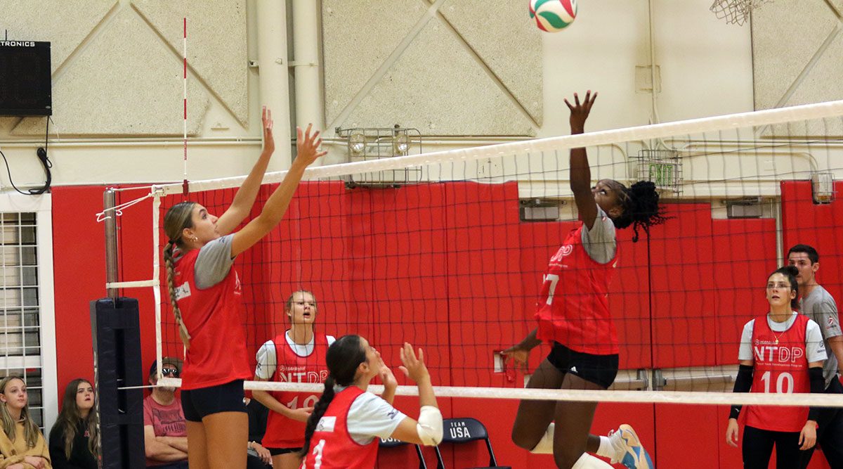 Two girls volleyball players at the net. One is hitting and the other is blocking. Other players and a coach watch