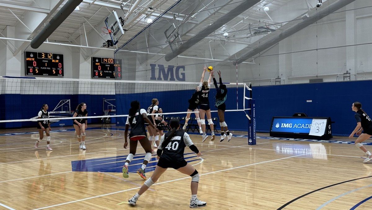 A far shot of a volleyball court with two teams playing. Two girls are up for a block