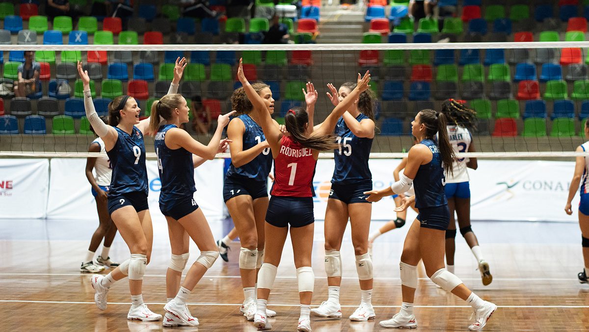 Six women on the volleyball court raise their hands and huddle